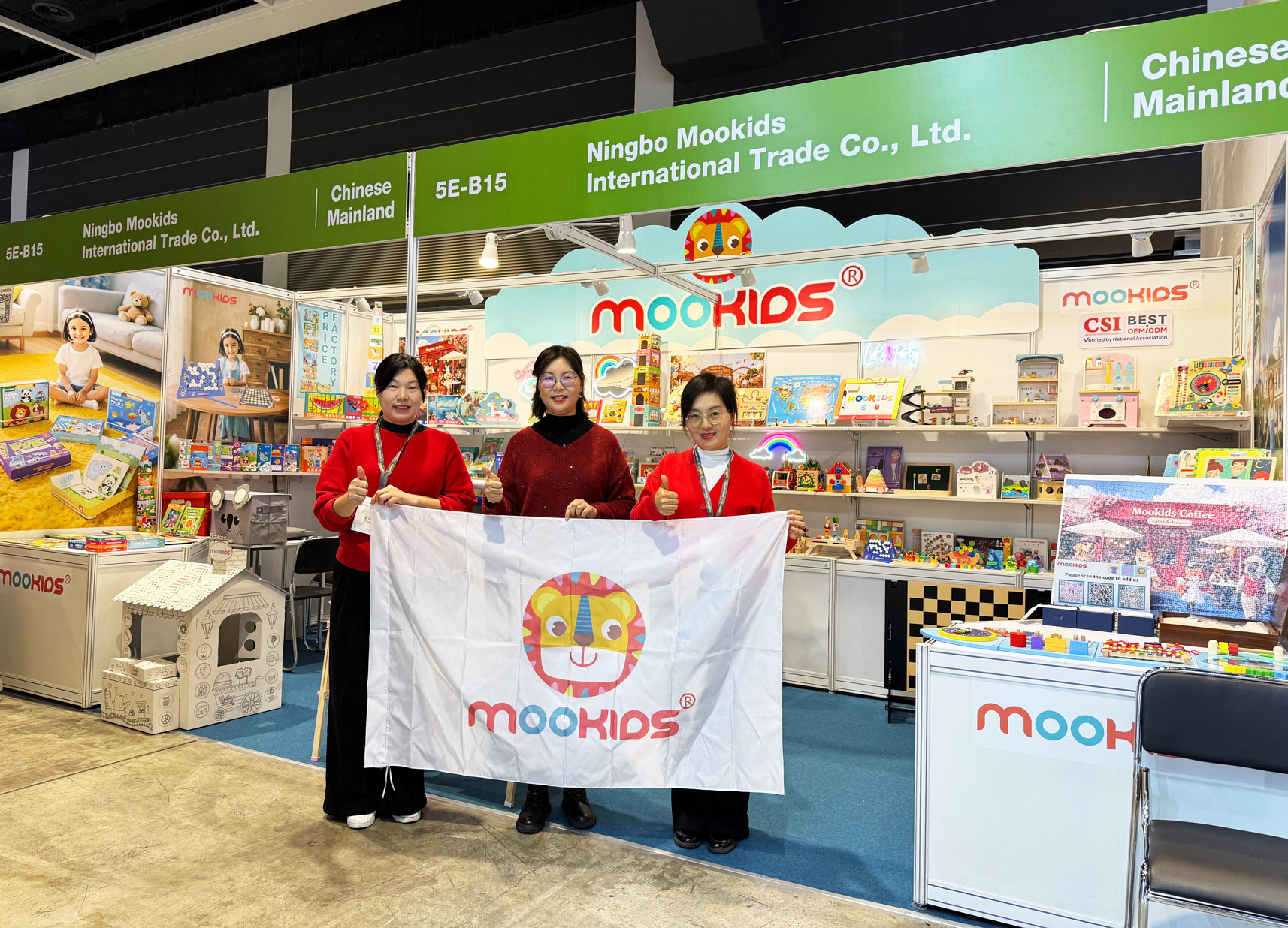 Three team members from Ningbo Mookids International Trade Co., Ltd. hold the Mookids brand banner at Booth 5E-B15 during the Hong Kong Toys & Games Fair, with a vibrant display of eco-friendly wooden toys and educational playthings in the background.