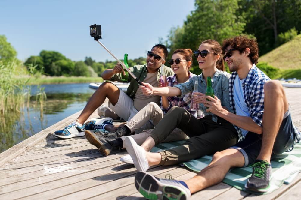 Several People Sit by the Lake and Take Group Photos with an Action Camera Mounted on a Selfie Stick