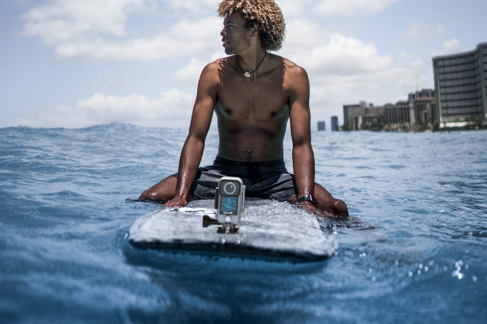 A Person Sits on a Surfboard on the Sea, and an SJCAM C300 Action Camera with Waterproof Case Mounted at the Front of the Surfboard