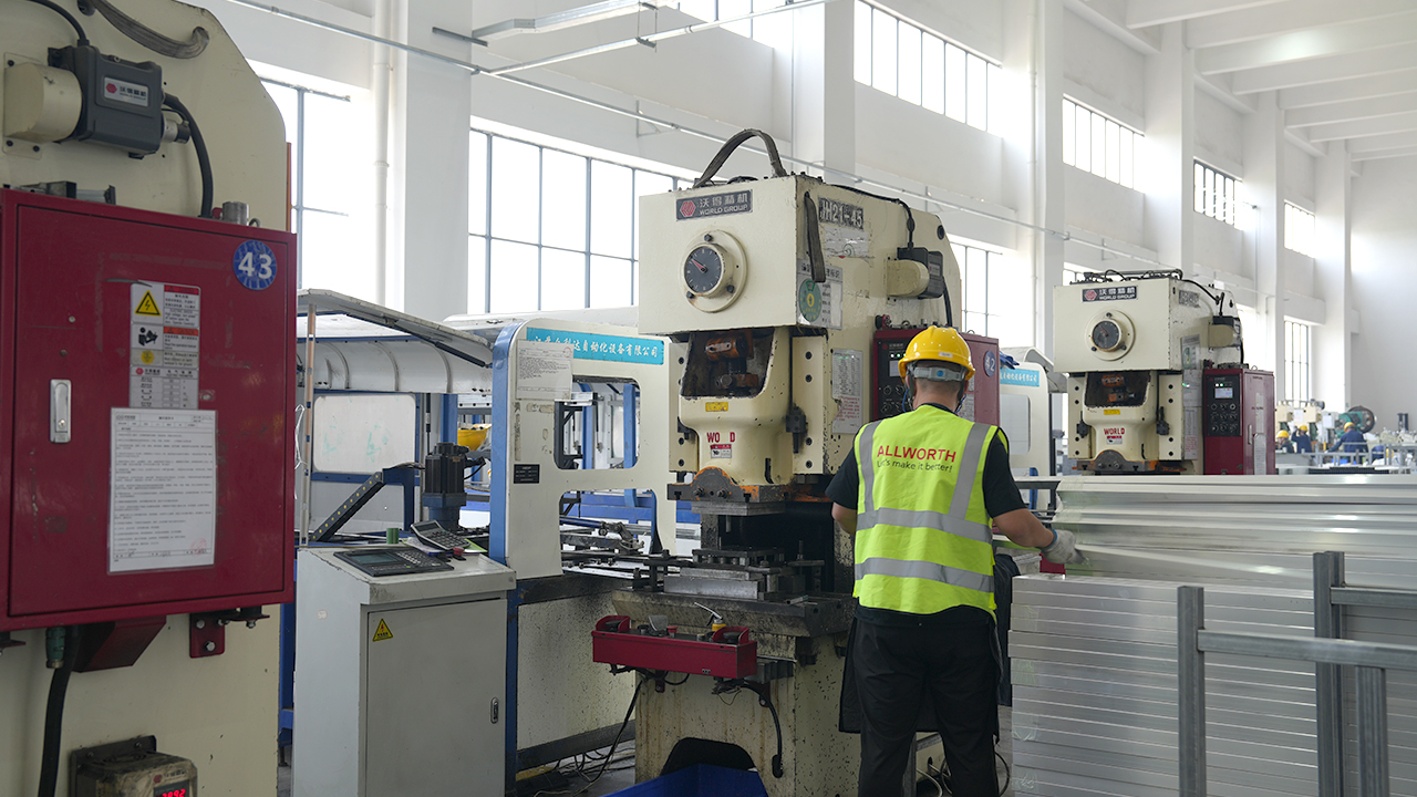 Worker in an ALLWORTH vest handling metal components beside production equipment