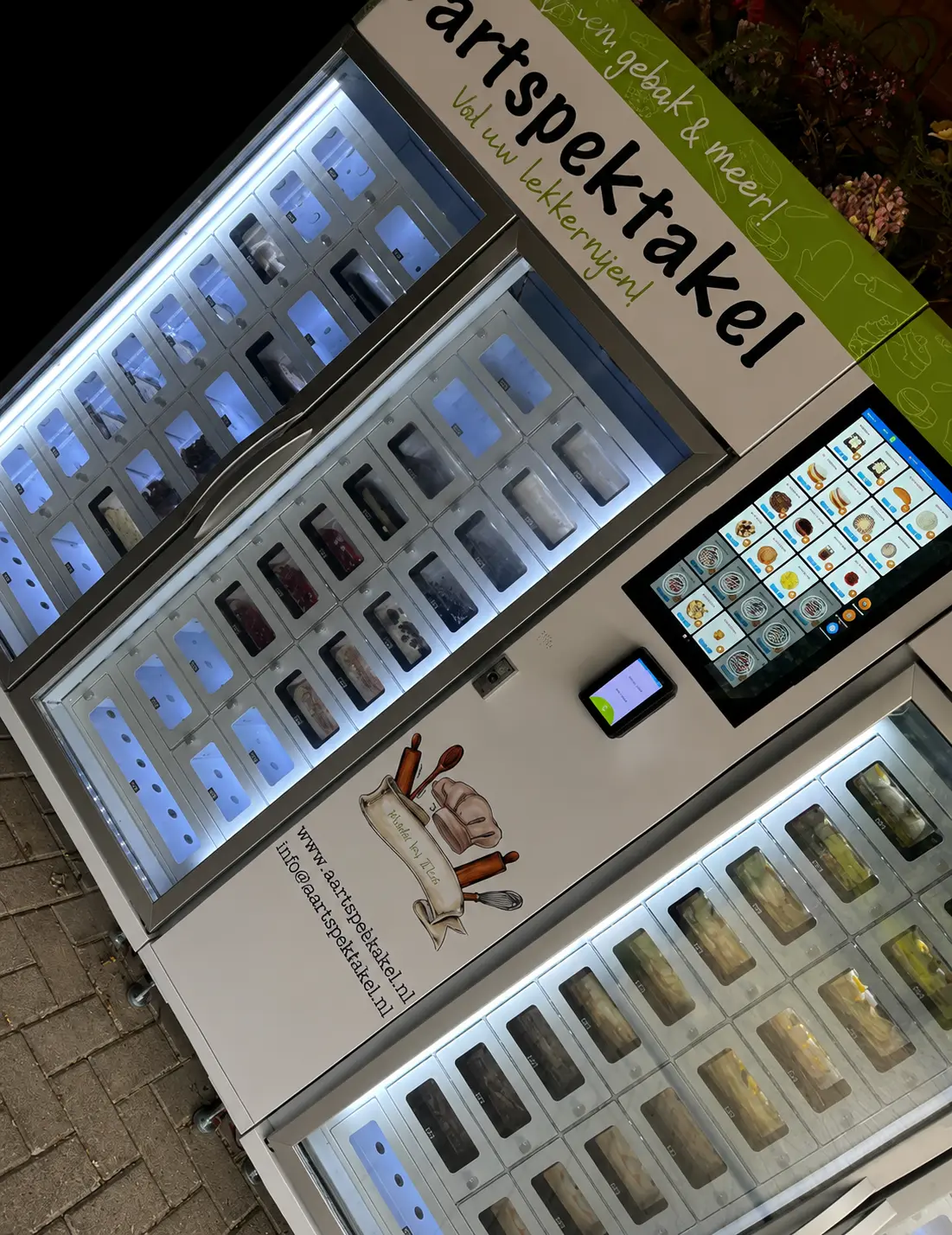 Illuminated outdoor pastry vending machine at night featuring multiple glass-door compartments, a touchscreen ordering panel, and a card payment terminal on a white and green cabinet.