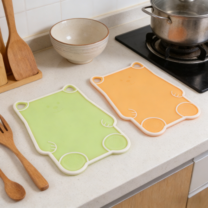 Two cute bear-shaped kitchen cutting boards (green and orange) with white trim, displayed on a light-colored kitchen countertop alongside utensils, a bowl, and a stove, highlighting their playful design and practical use in food preparation.