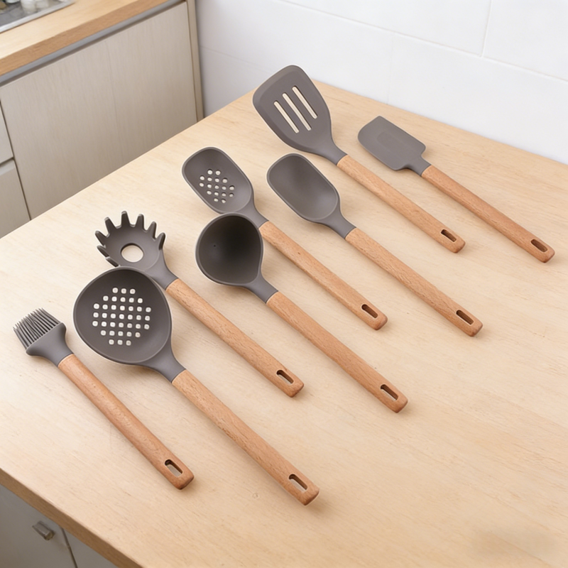8-piece gray silicone kitchen utensil set with wooden handles, neatly arranged on a light wood kitchen countertop, including a slotted turner, small spatula, slotted spoon, solid spoon, pasta fork, ladle, skimmer, and basting brush.