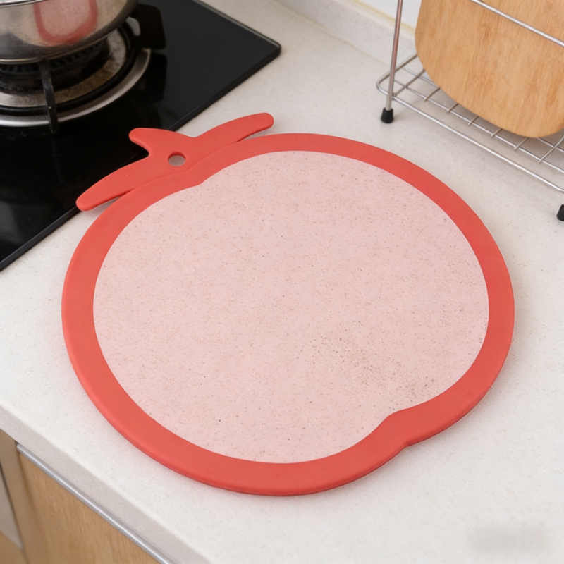 Tomato-shaped kitchen cutting board with a bright red border and light pink wheat-straw surface, featuring a stem-style hanging hole, displayed on a clean countertop next to a gas stove in a modern kitchen.