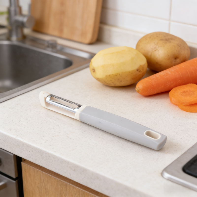 Gray and white serrated vegetable peeler with stainless steel blade, placed on a light kitchen countertop alongside a peeled potato, whole potato, and fresh carrots, with a sink in the background.