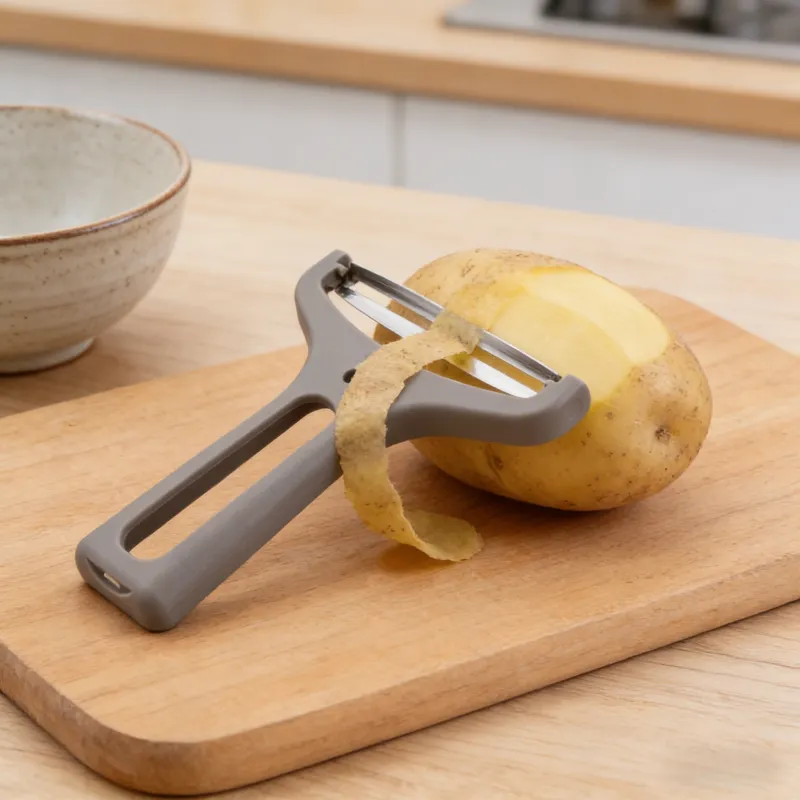 Gray Y-shaped vegetable peeler with stainless steel blade peeling a potato on a wooden cutting board, with potato peel attached, in a warm kitchen setting.