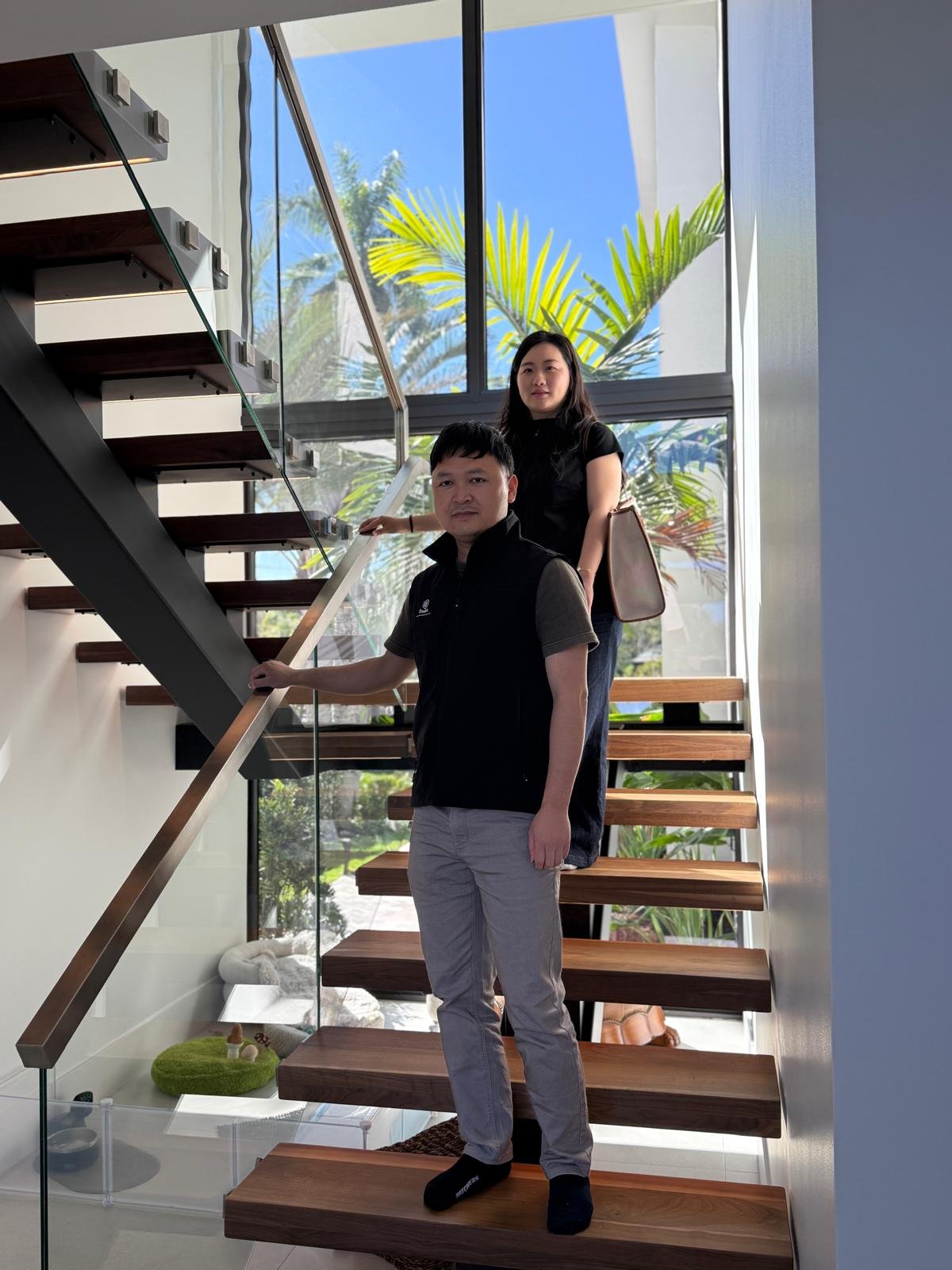 Two people standing on a floating staircase with glass railing and palms beyond the window