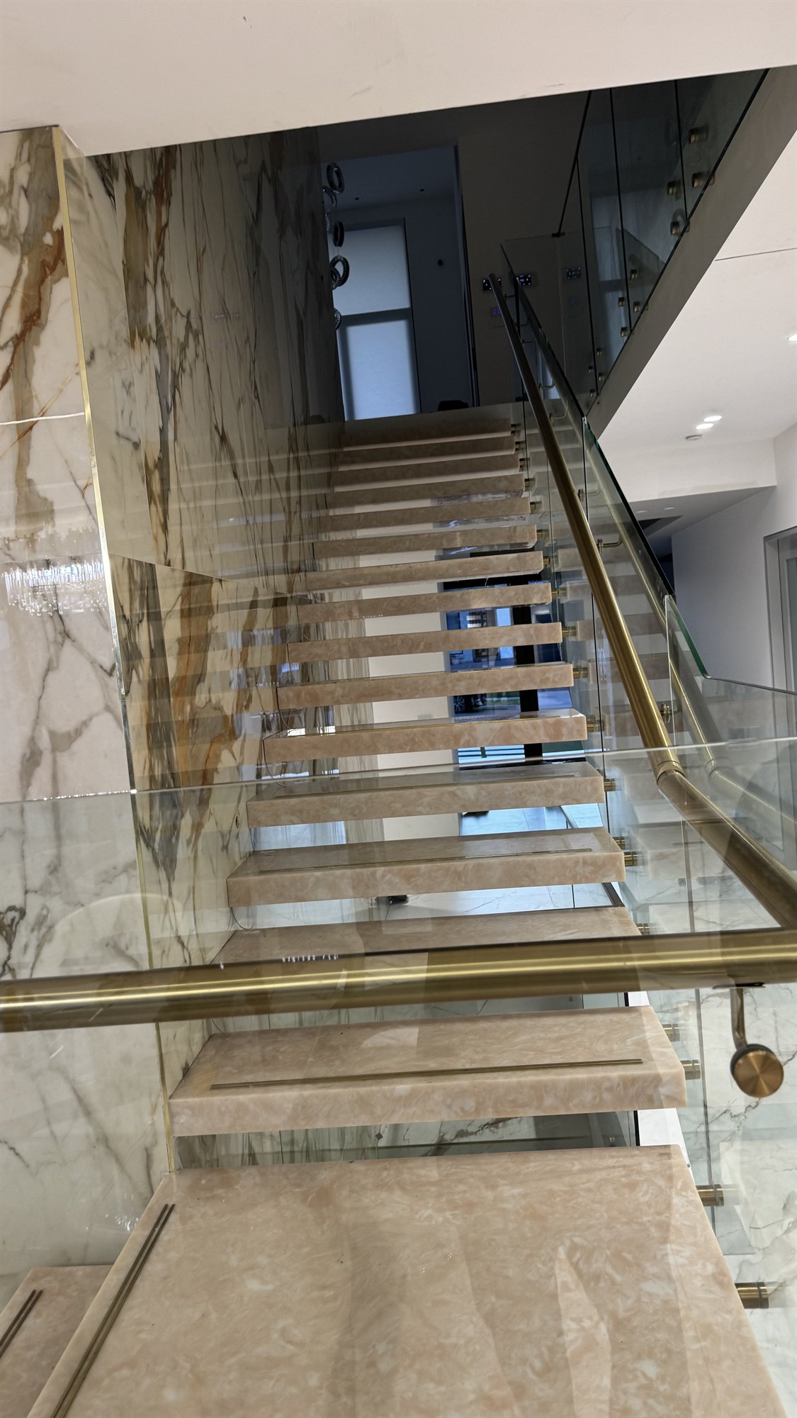 Looking up a pale marble floating staircase with glass panels and a brass handrail against a stone wall