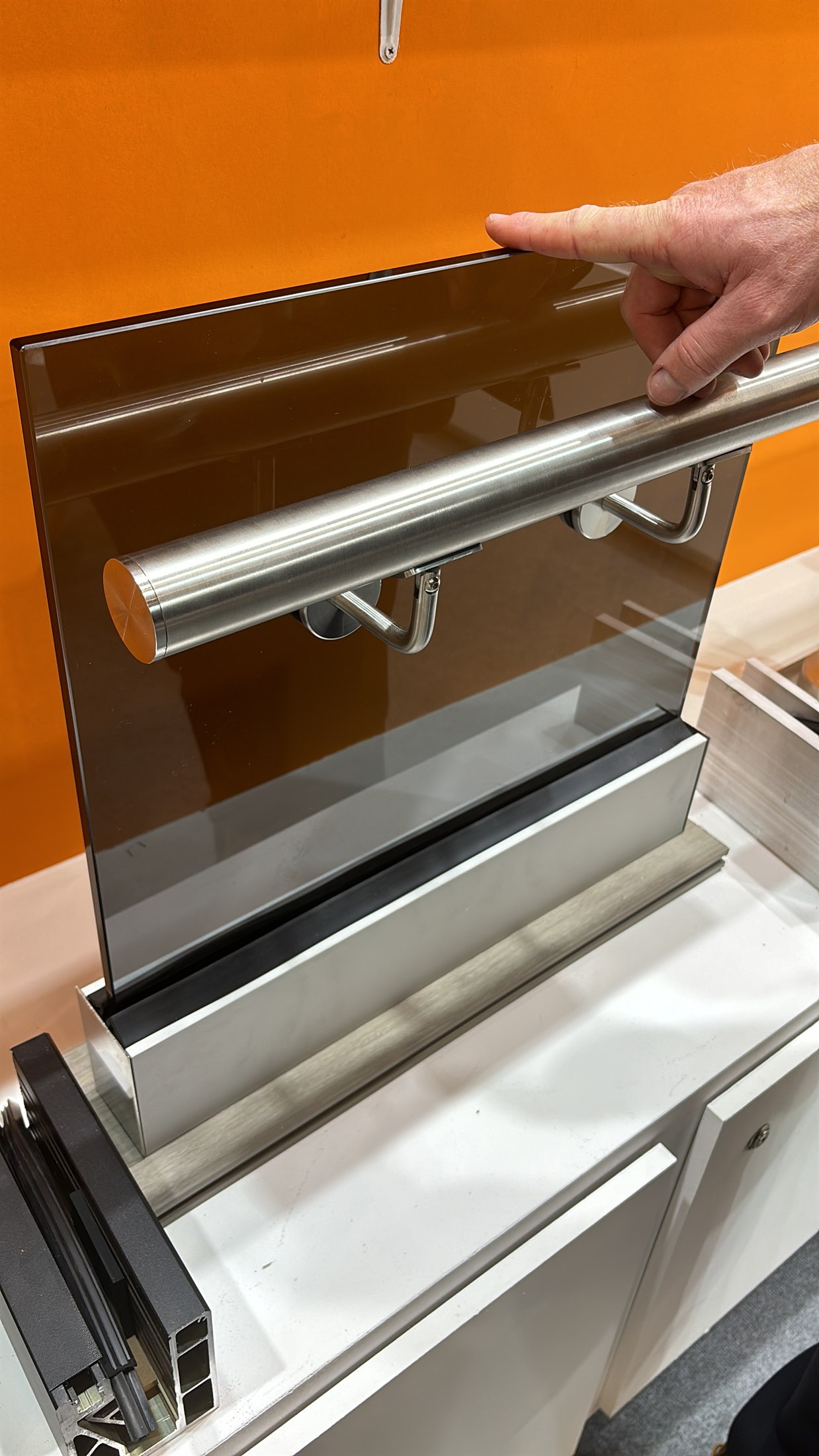 Bronze-tinted glass railing panel in a metal base channel with a round stainless handrail, a hand at the top edge