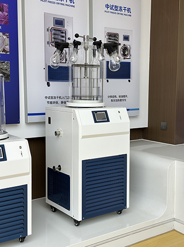Front showroom view of a manifold stoppering laboratory freeze dryer with glass flasks attached, displayed in front of pilot freeze-drying machine posters.