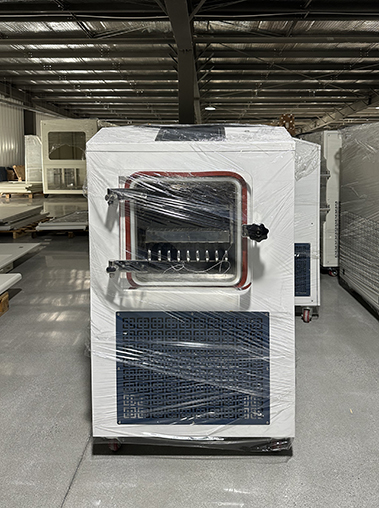 Right side angled view of a white vacuum freeze dryer machine wrapped in plastic film inside a warehouse facility.
