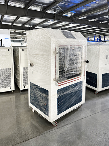 Left perspective view of a white pilot vacuum freeze dryer wrapped in protective plastic film inside a factory workshop.