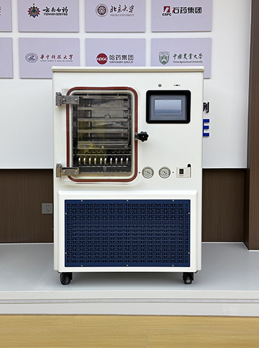 Front straight-on view of a white vacuum freeze dryer machine showcased on a display platform against a corporate partner wall.