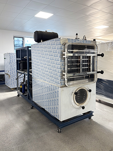 Side perspective real shot of a laboratory freeze dryer unit on wheels with blue protective film inside a workshop facility.