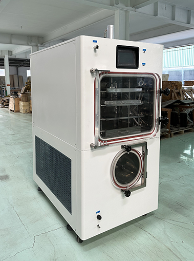 Three-quarter left view real shot of a white vertical pilot vacuum freeze dryer unit inside a manufacturing workshop, showing its deep side panel and mesh vent.