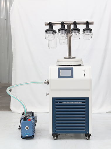 Angled workshop real shot of a T-frame vacuum freeze dryer connected to a rotary vane vacuum pump via a reinforced transparent steel wire hose.